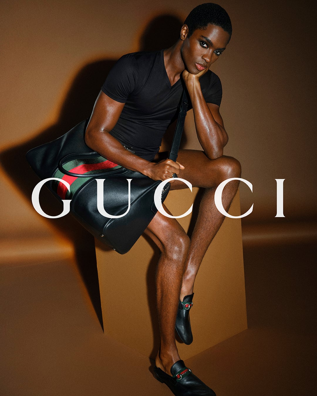 Alton Mason seated against a warm brown backdrop printed with the white Gucci logo in a fitted black short-sleeve tee, a large black leather duffel banded in the red-and-green Web stripe held over the lap, black GG Horsebit loafers worn barefoot