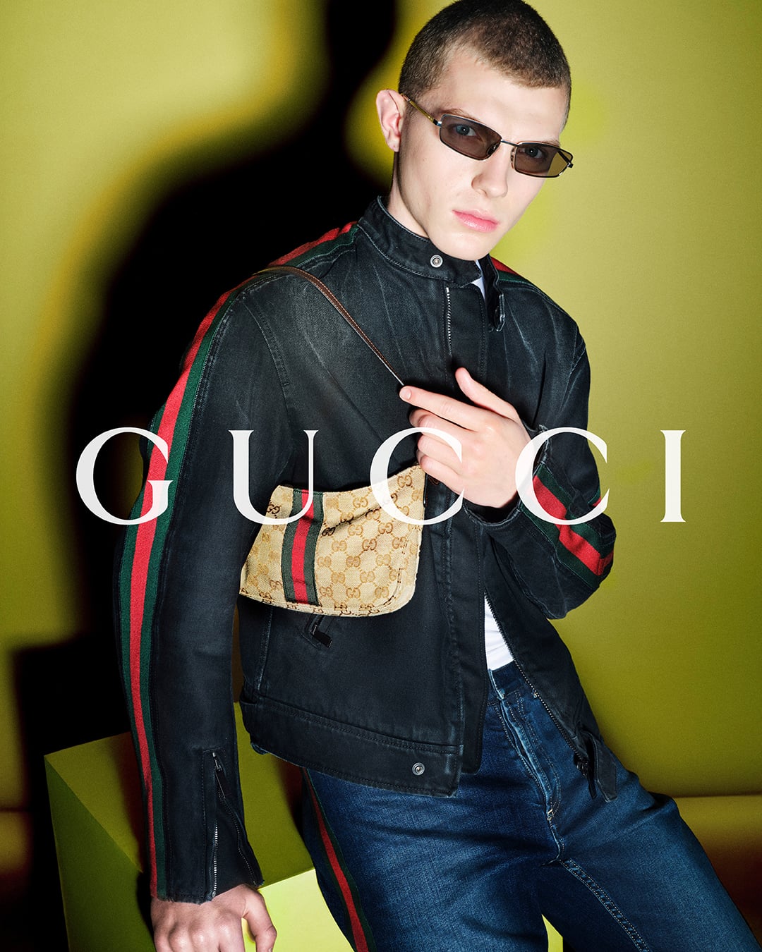 Bede Harper close up against a chartreuse backdrop printed with the white Gucci logo, wearing a black denim moto jacket striped in red and green down the sleeves and a small GG-monogram canvas pouch with Web stripe over the shoulder, slim rectangular sunglasses