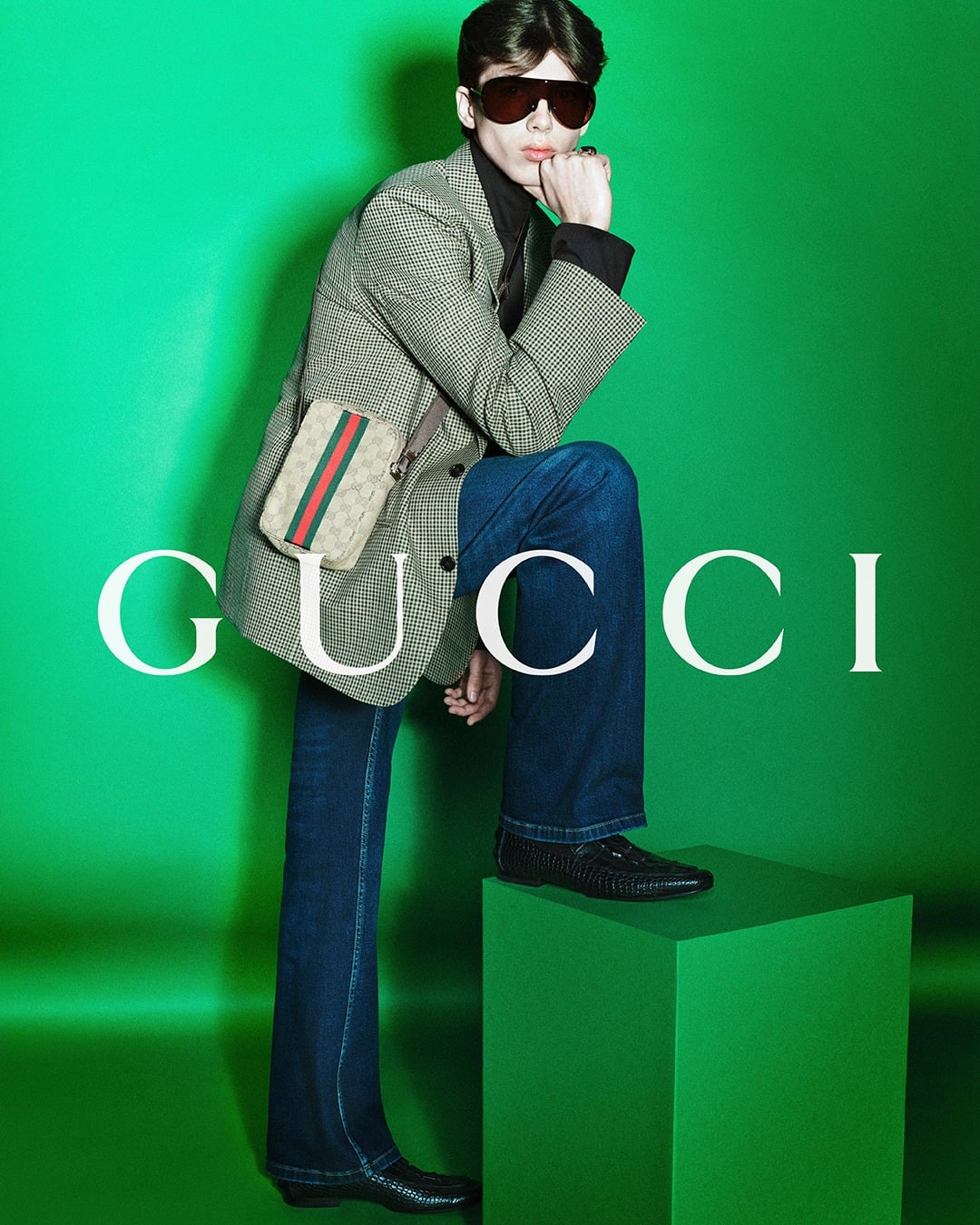 Ben Eckstein standing against a vivid green backdrop printed with the white Gucci logo in an oversized houndstooth blazer over a black turtleneck and dark wash flared jeans, a small GG-monogram canvas pouch with red-and-green Web stripe in the breast pocket, oversized amber-tinted shield sunglasses, one foot propped on a green plinth