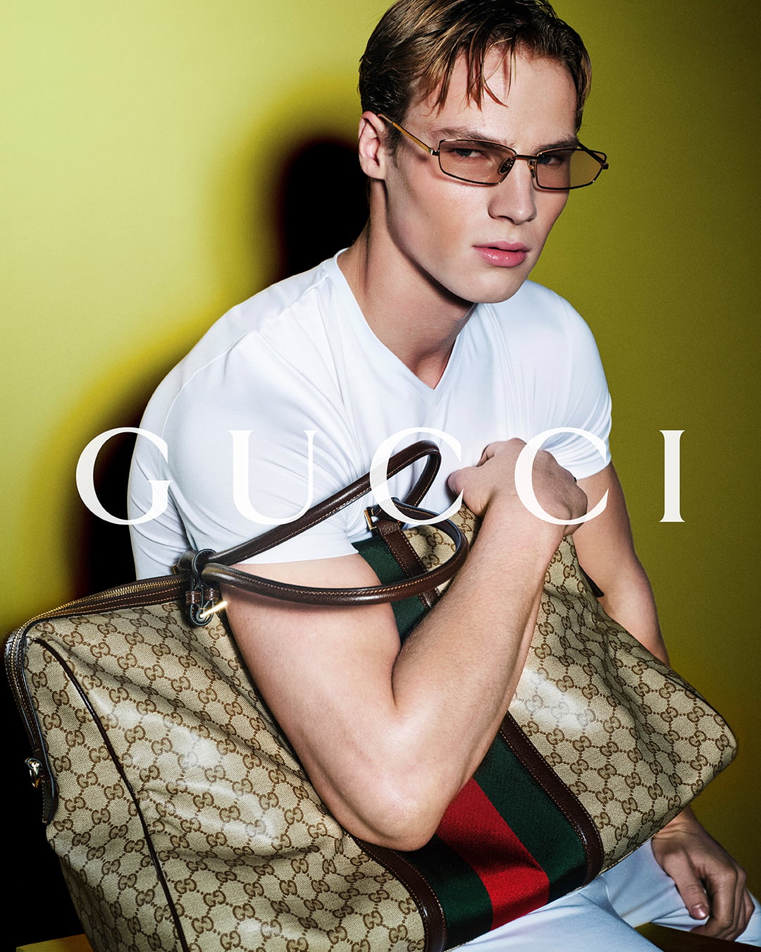 Henry Rank with damp blond hair and amber-tinted rectangular sunglasses, wearing a fitted white short-sleeve V-neck tee and pale trousers against a chartreuse yellow backdrop printed with the white Gucci logo, holding a large GG-monogram canvas tote bordered with brown leather and the red-and-green Web stripe close to the chest