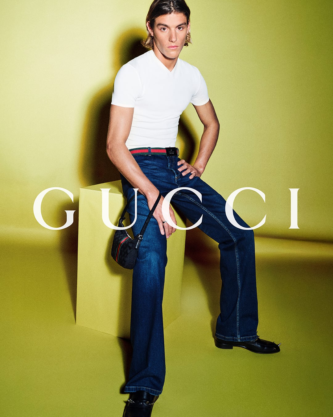 Vinicius Vieira with shoulder-length brown hair perched on a yellow plinth in a fitted white short-sleeve V-neck tee and dark wash flared jeans cinched with a red-and-green Web stripe belt, a compact suede Web-stripe shoulder bag hanging at the hip against a chartreuse backdrop printed with the white Gucci logo