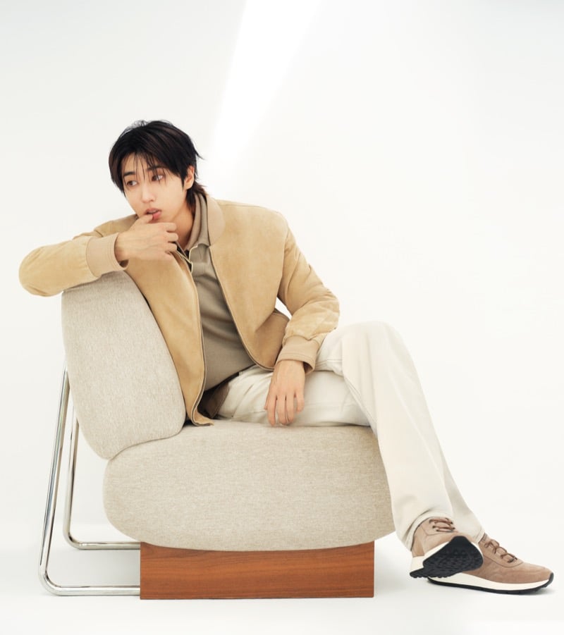 Han Jisung reclines on a cream upholstered armchair with a chrome and wood frame in a tan suede bomber, taupe polo, cream pants, and brown suede Tod's sneakers