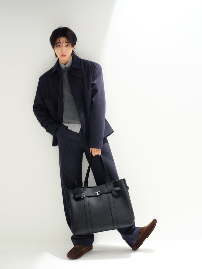 Han Jisung leans against a white wall in a wool overshirt and wide-leg trousers layered over a gray sweater and button-down shirt, holding a black leather Tod's T tote with brown suede Gommino loafers