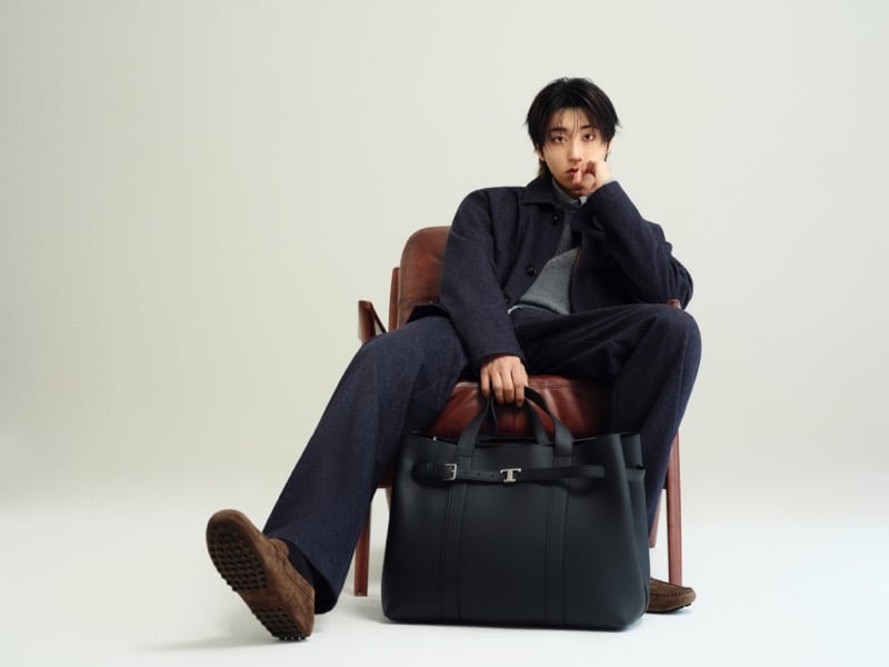 Han Jisung sits in a leather and wood armchair in a wool jacket, gray sweater, and brown suede Gommino loafers with a black leather Tod's T tote resting at his feet