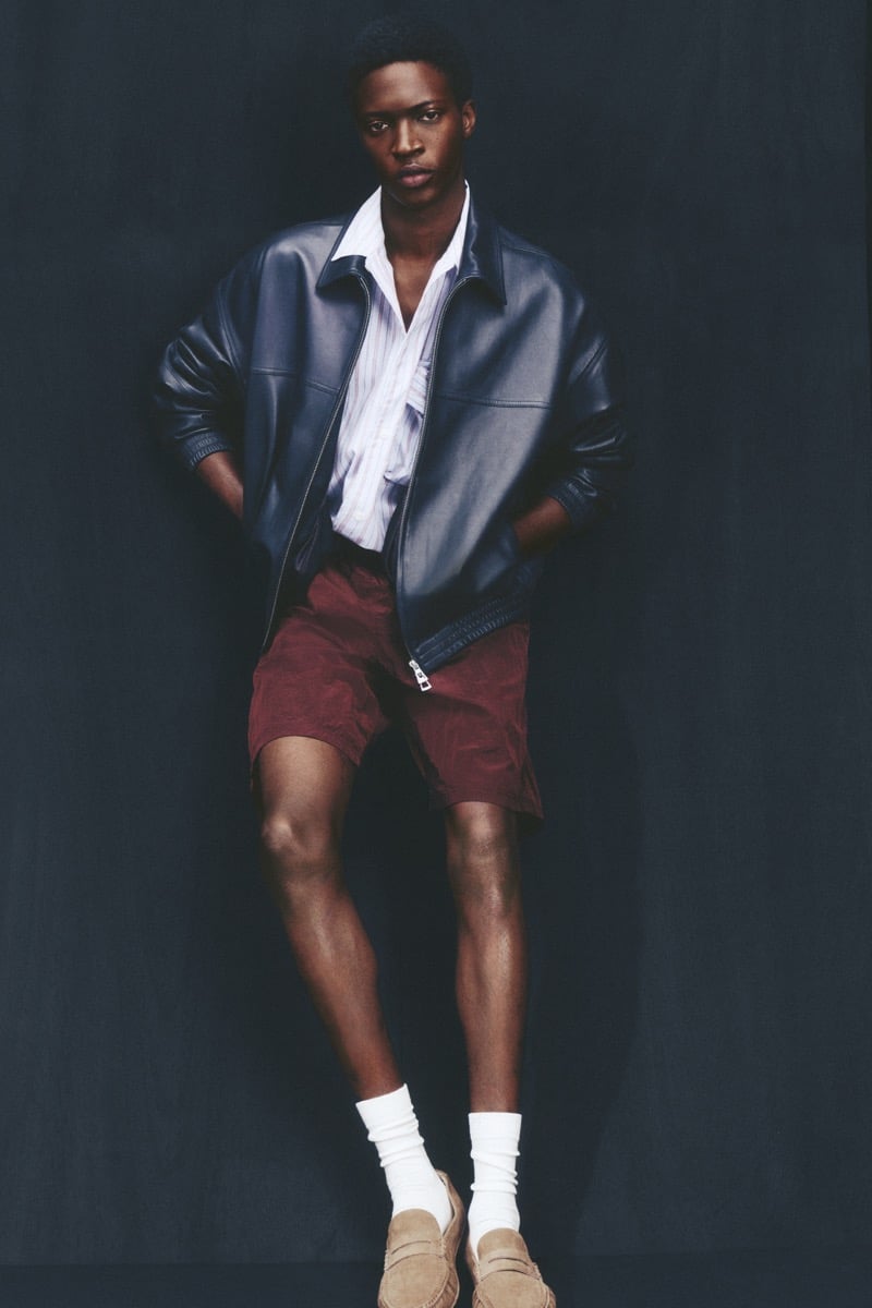 Limamu Mbaye wears a dark leather zip-front blouson over a thin-striped shirt with burgundy shorts, slouched white socks, and tan suede driving loafers against a deep backdrop