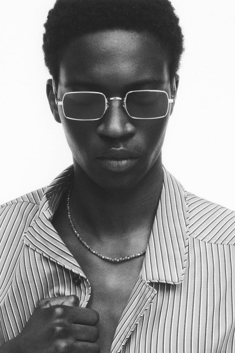 Close-up of Limamu Mbaye in slim square wire-rim sunglasses and a beaded necklace, wearing an open thin-striped collared shirt