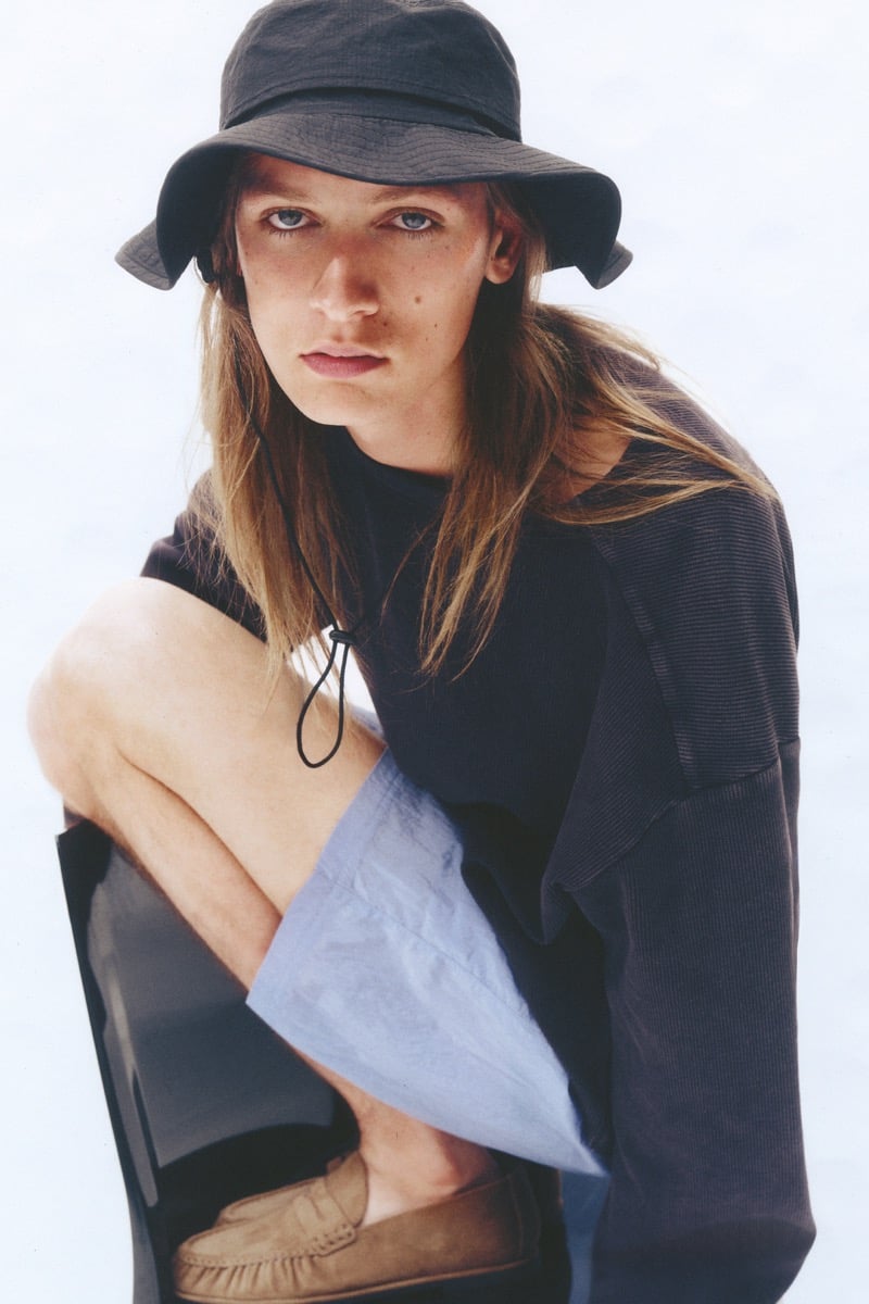 Jesse Rinderknecht wears a floppy dark bucket hat with a chin cord and a textured top, accented by pale blue shorts with tan suede loafers