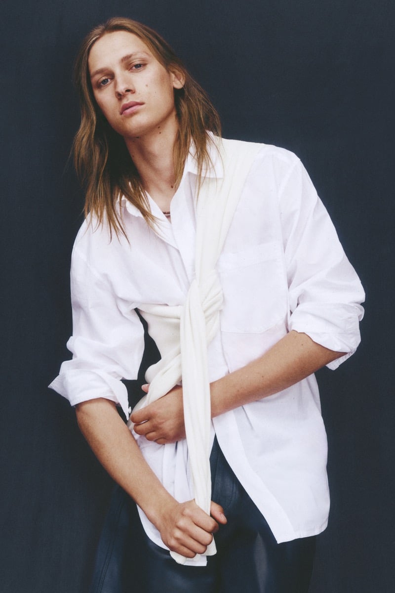 Jesse Rinderknecht wears a roomy crisp white button-up shirt with a soft knit tied around the shoulder and leather shorts against a dark backdrop