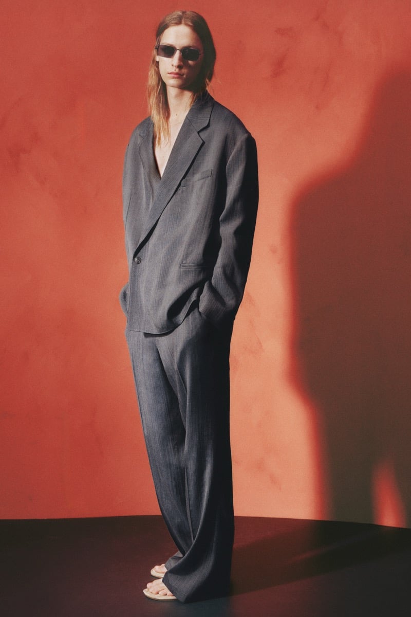 Jesse Rinderknecht wears slim sunglasses with a relaxed charcoal double-breasted suit worn open over a bare chest, paired with pale sandals against a terracotta wall