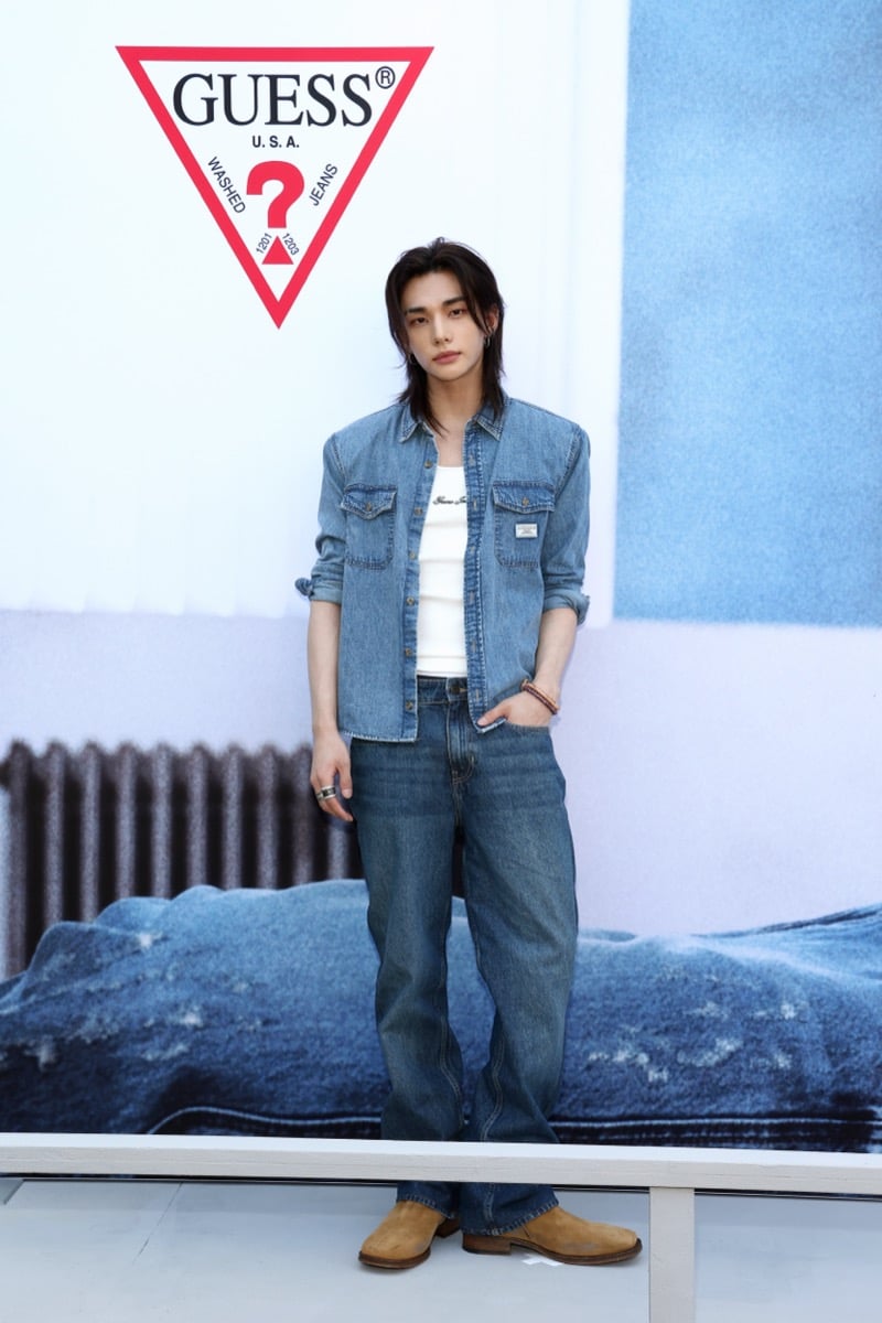 Hyunjin standing in front of a large GUESS triangle logo backdrop at the Melrose flagship in a blue denim shirt over a white ribbed tank with loose blue jeans and tan suede western boots