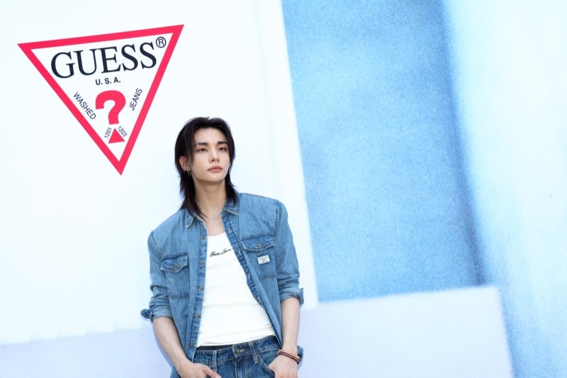 Hyunjin leaning against a wall printed with the GUESS triangle logo in a blue denim shirt over a white ribbed tank and dark jeans