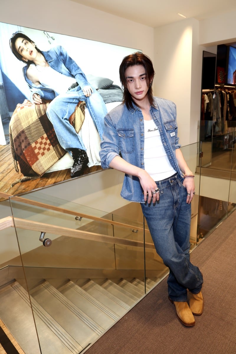 Hyunjin posing beside a large in-store print of his GUESS campaign image, wearing a blue denim shirt over a white ribbed tank, loose blue jeans, and tan suede boots