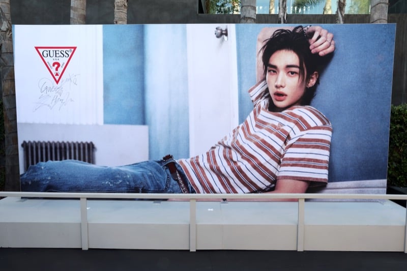 Large outdoor GUESS campaign panel of Hyunjin reclining in a red striped t-shirt and washed jeans, signed with his autograph