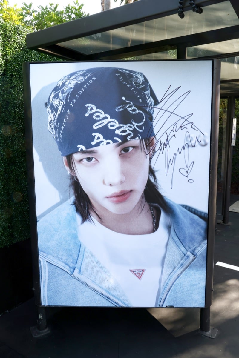 Bus shelter advertisement showing Hyunjin in a paisley bandana and a light denim jacket over a white t-shirt with a GUESS triangle logo, signed in marker