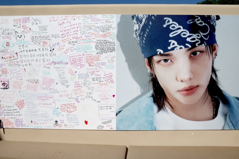 Fan message wall covered in handwritten notes to Hyunjin next to a campaign panel of him in a navy paisley bandana