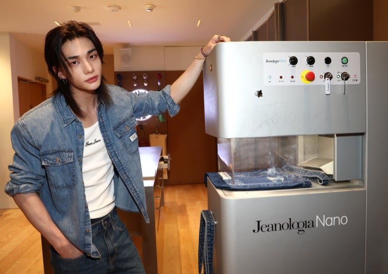 Hyunjin posing beside a Jeanologia Nano denim laser personalization machine at the GUESS pop-up in a blue denim shirt and white tank