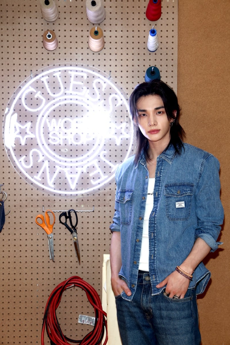 Hyunjin standing in front of a pegboard with a GUESS Jeans neon sign and thread spools at the denim customization station