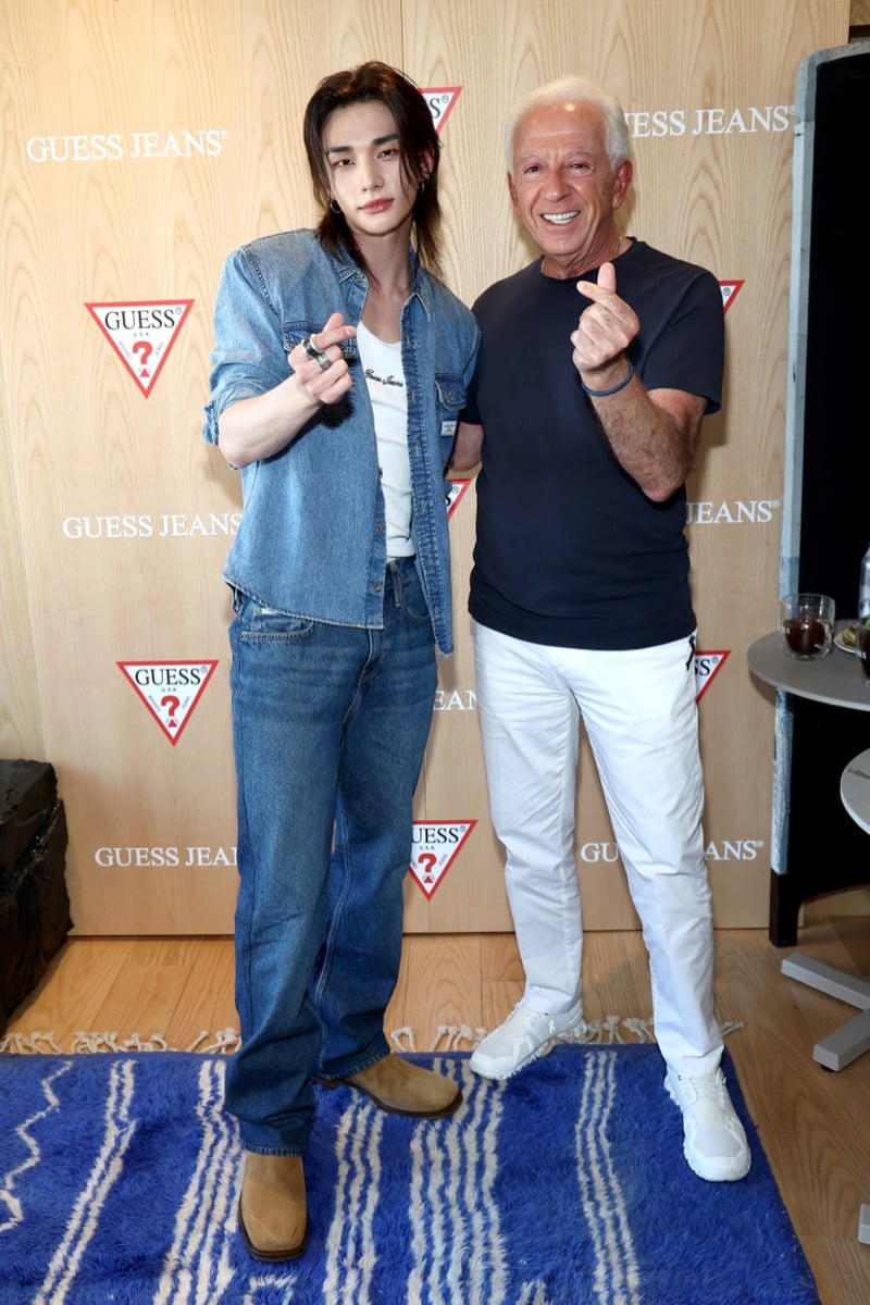 Hyunjin and Paul Marciano making finger heart gestures together in front of the GUESS Jeans backdrop