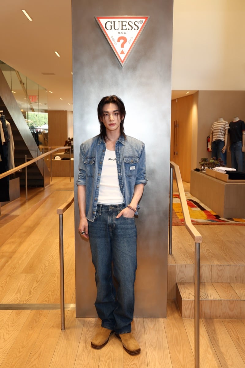 Full-length shot of Hyunjin standing in front of a silver pillar topped with the GUESS triangle logo inside the Melrose flagship