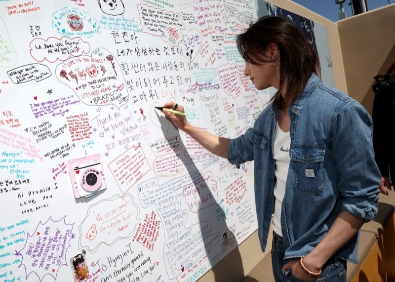 Hyunjin signing a fan message wall filled with handwritten notes during the GUESS Melrose pop-up