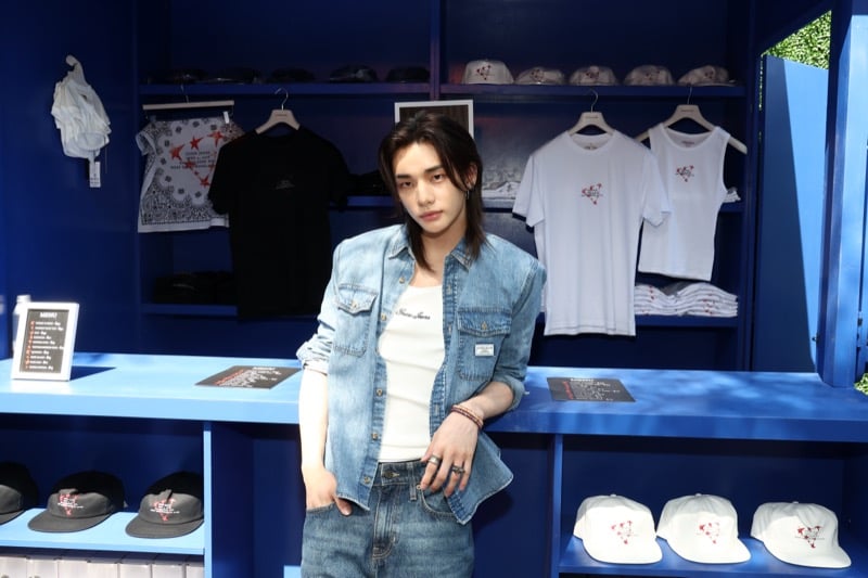 Hyunjin standing in front of a blue merchandise wall displaying exclusive GUESS pop-up t-shirts and caps, wearing a blue denim shirt over a white ribbed tank and dark jeans