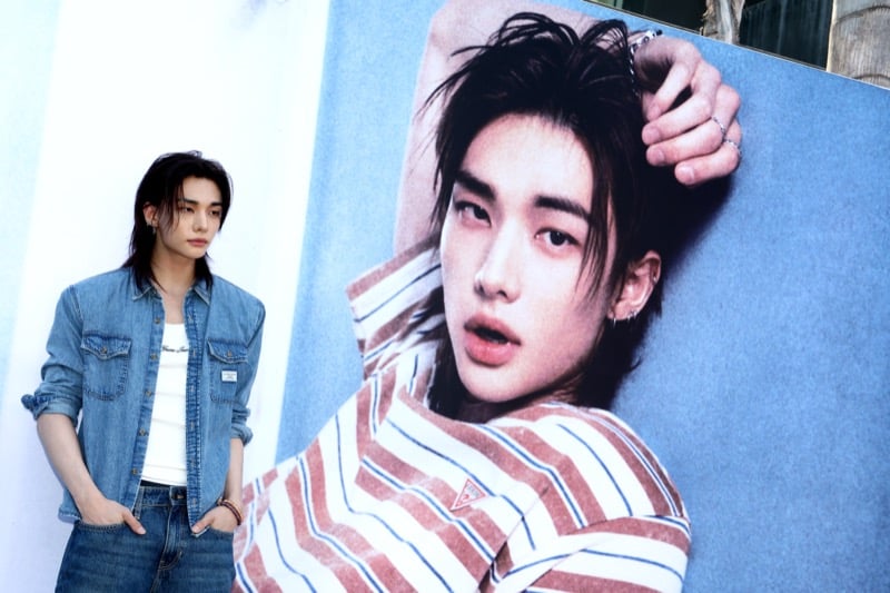 Hyunjin standing beside a large outdoor campaign poster of himself in a red striped t-shirt and bandana, wearing a blue denim shirt over a white ribbed tank