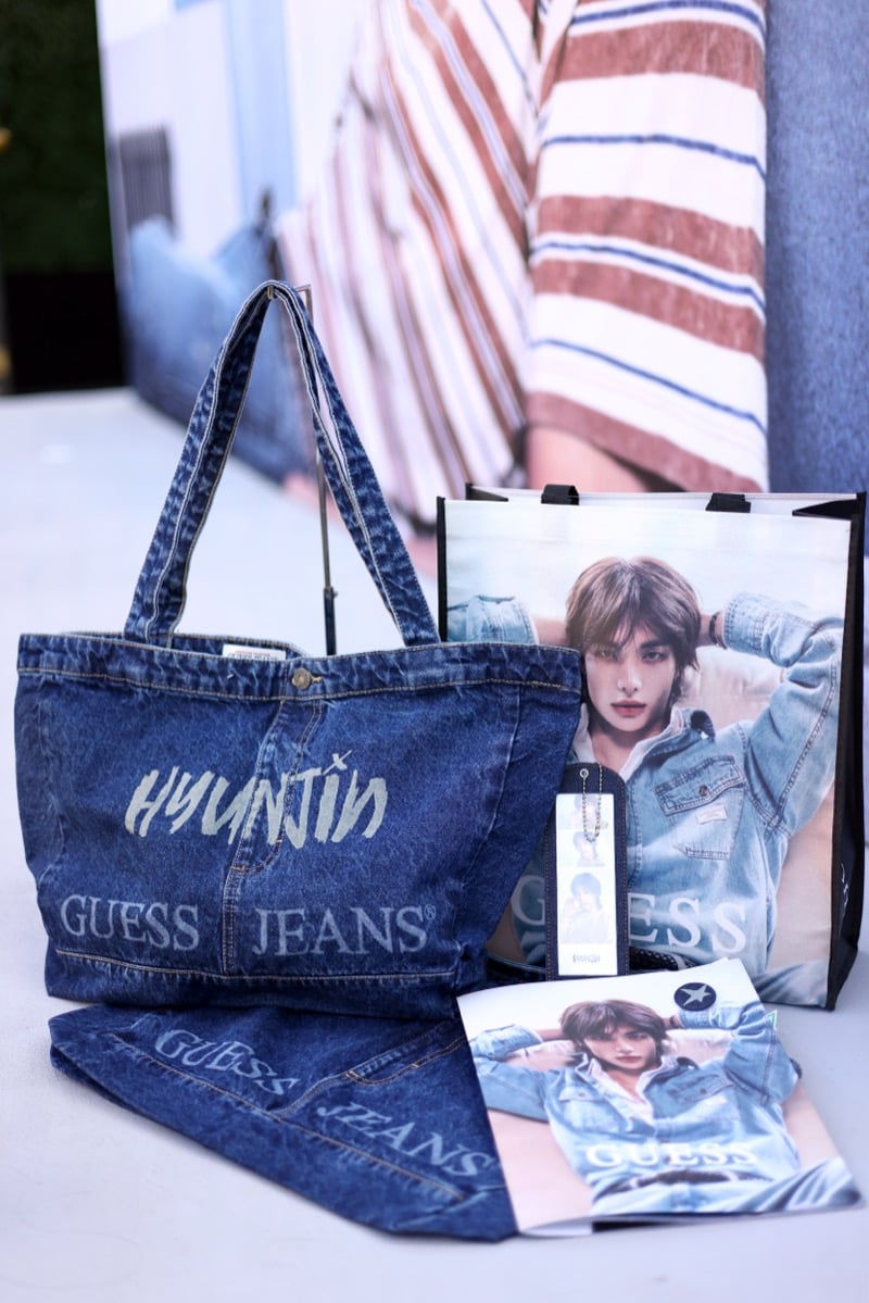Limited-edition GUESS Jeans denim tote bag printed with Hyunjin's name beside a campaign-printed shopping bag and exclusive pop-up giveaways