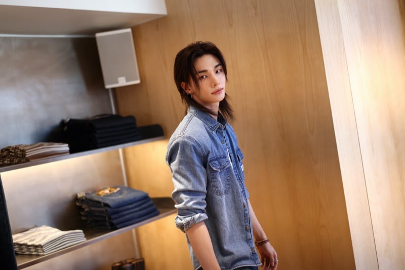 Hyunjin turning to look at the camera between stacked denim shelves inside the GUESS Melrose flagship in a blue denim shirt