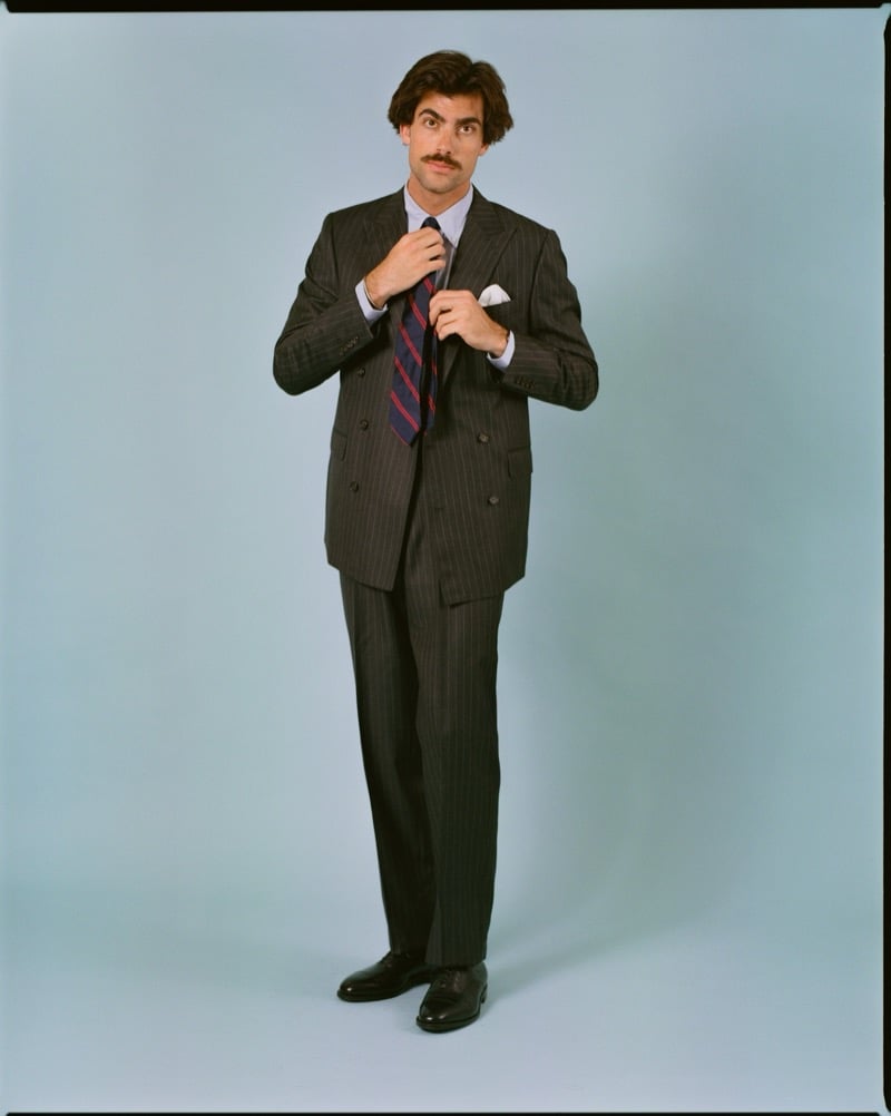Victor Netland adjusts a striped repp tie while modeling the J. Press Mayfair double-breasted suit in a dark chalkstripe with a light blue dress shirt
