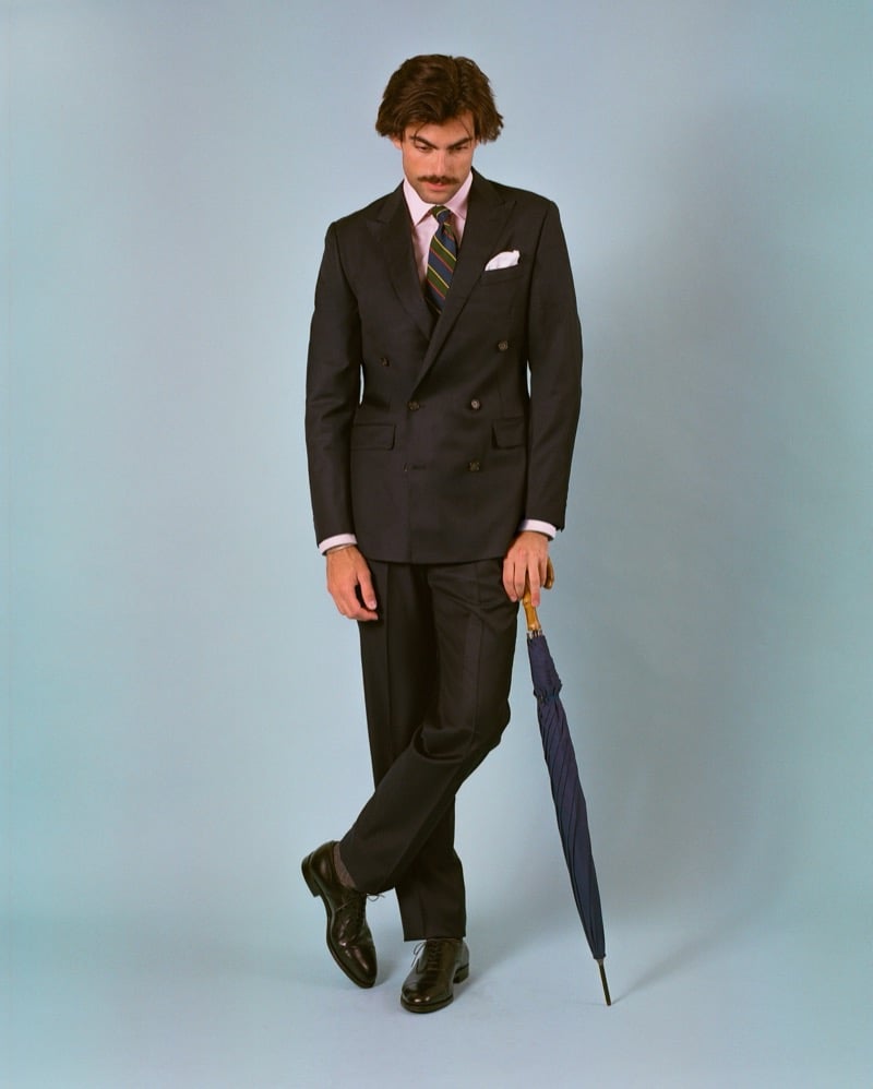 Victor Netland holds a wood-handled umbrella in the J. Press Mayfair double-breasted suit, paired with a pink shirt, a multi-stripe tie, and dark derby shoes