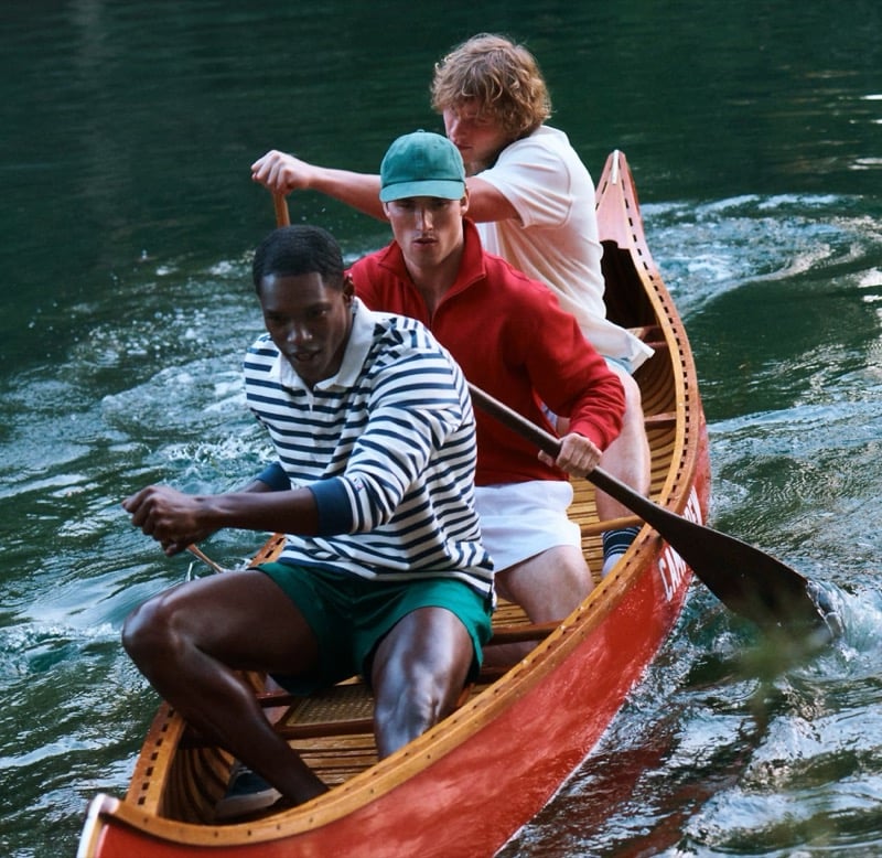 Models Ronald Epps, Jack Lumsden, and Anthony Foster paddling a wooden red canoe on the lake for J.Crew's Camp Crew summer 2026 campaign, wearing a striped rugby shirt with green shorts, a red knit polo with a green cap, and a white crewneck sweater