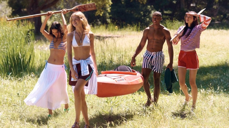 Three female cast members and a shirtless Ebeneze Brown carry an orange kayak and wooden paddles through a sunlit grassy field for J.Crew's Camp Crew summer 2026 campaign, layering swimwear with skirts, shorts, and a striped tee