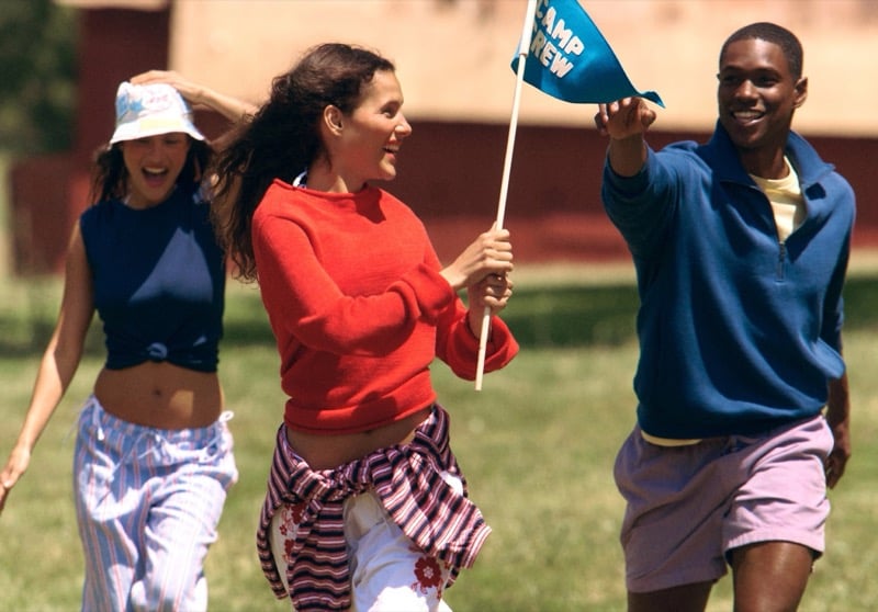 Two women and Ronald Epps running across a camp lawn for J.Crew Camp Crew summer 2026, one woman holding a blue Camp Crew pennant flag while the Ronald in a blue quarter-zip cheers behind her