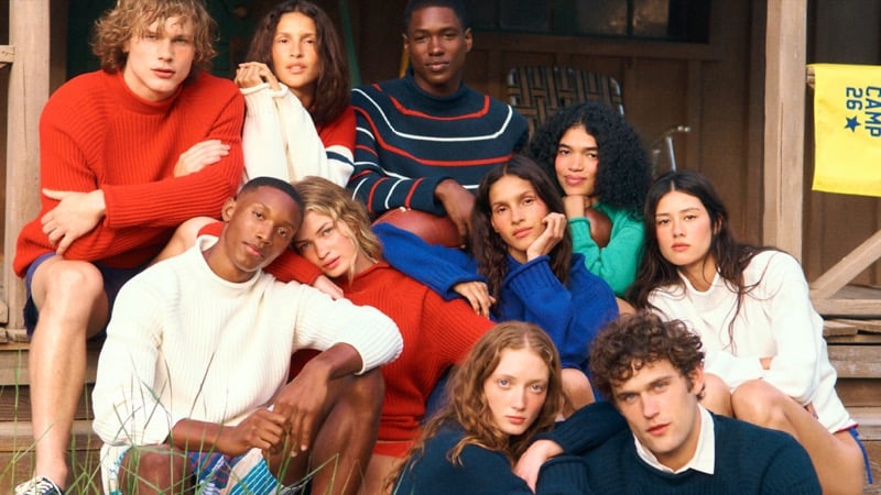 J.Crew Camp Crew summer 2026 group portrait on cabin steps featuring the cast in rugby knits, striped rollnecks, and chunky pullovers, with a yellow Camp 26 pennant hanging in the background