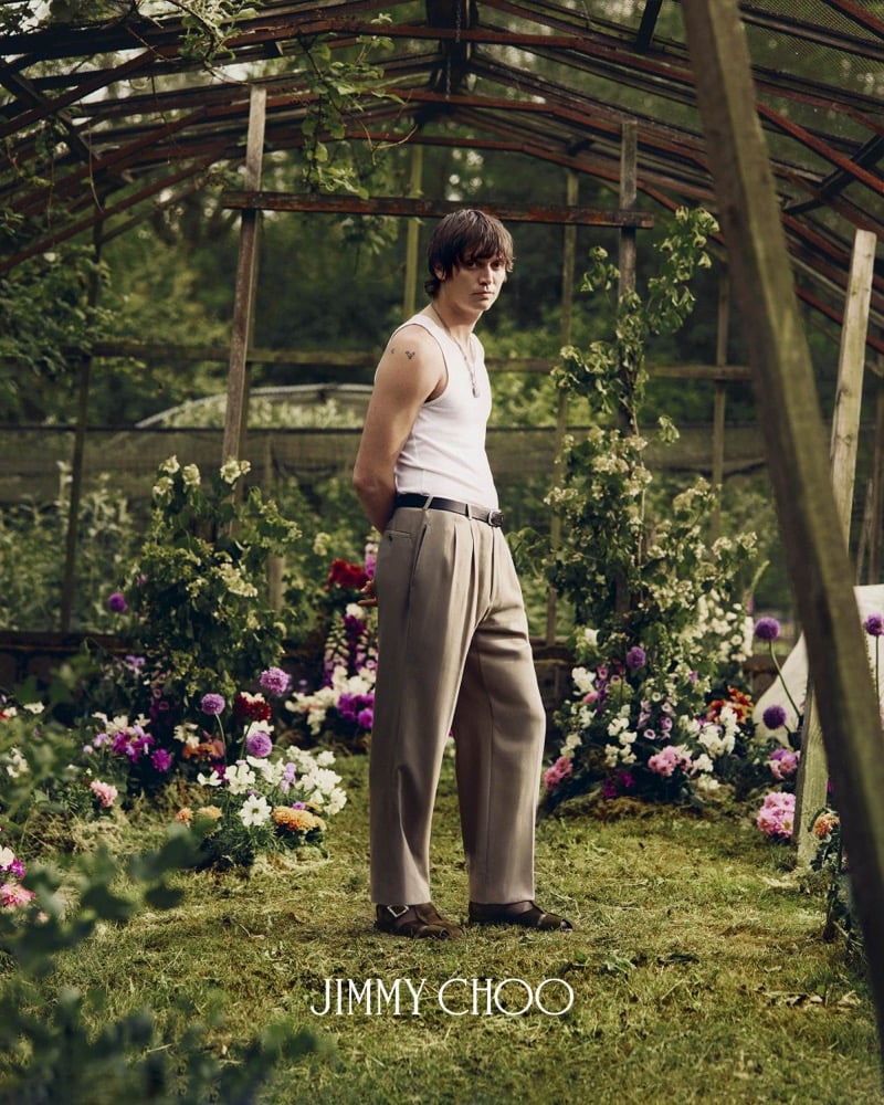 Alfie Nickerson standing inside a wood-frame greenhouse with cultivated flower beds on either side, wearing a white ribbed cotton tank top and taupe pleated wide-leg trousers belted with a slim black leather strap, finished with brown leather buckled sandals