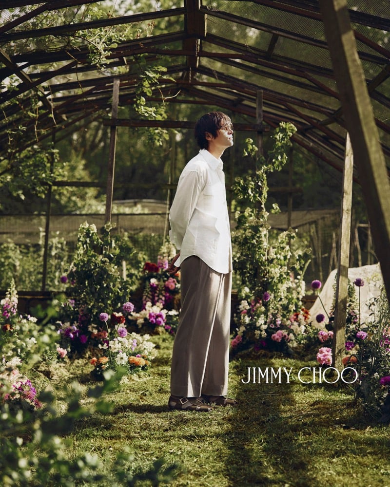Alfie Nickerson photographed in profile inside the greenhouse, wearing a loose white mandarin-collar linen shirt tucked into taupe wide-leg trousers, with brown leather buckled sandals on the grass below