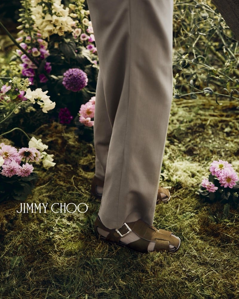 Close-up of Alfie Nickerson's feet in brown suede Jimmy Choo Meadow fisherman sandals with a polished silver buckle, worn beneath the hem of taupe wool trousers on a grass floor lined with pink dahlias and snapdragons