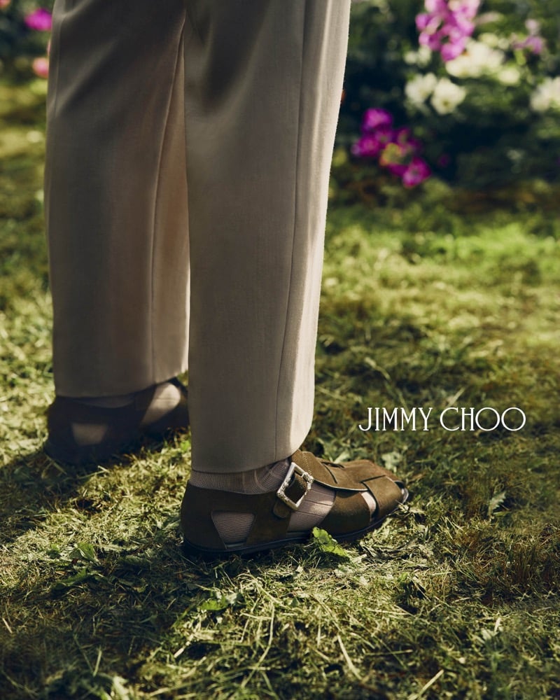 Tight overhead crop of Alfie Nickerson's feet stepping forward in brown suede Jimmy Choo Meadow sandals with a sculpted silver buckle, the taupe trousers breaking just above the leather straps