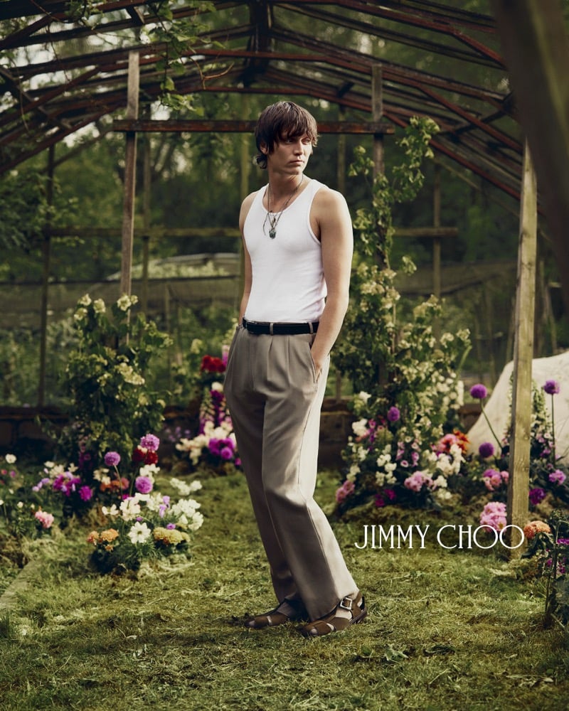 Alfie Nickerson posed full-length inside the greenhouse with hands at his pockets, in a white ribbed cotton tank, slim black leather belt, taupe pleated wide-leg trousers, and brown leather fisherman sandals, framed by beds of dahlias and roses