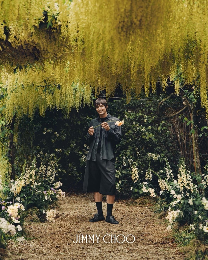 Alfie Nickerson smiling beneath a cascading laburnum archway in full yellow bloom, wearing a gray utility-pocket jacket over a matching mandarin shirt, black tailored shorts, ribbed black knee socks, and dark woven leather Jimmy Choo Eliot Slippers