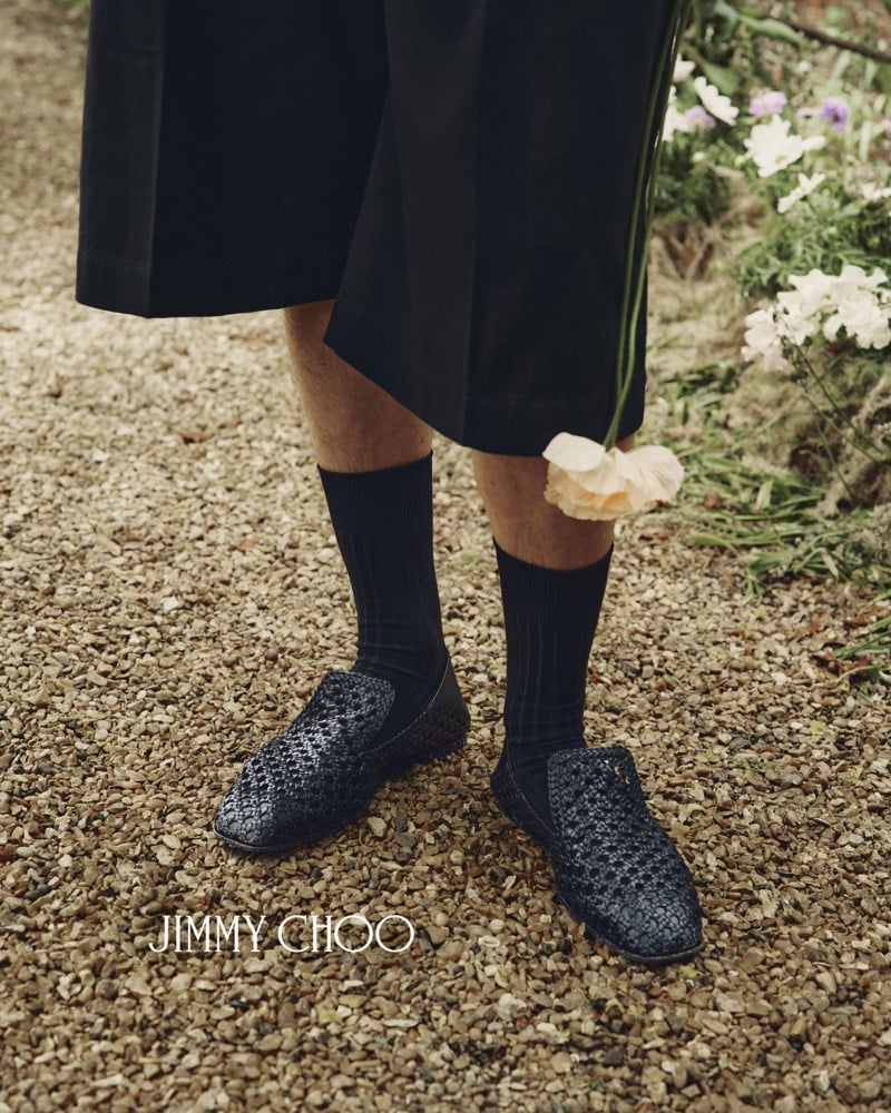 Close-up of Alfie Nickerson's feet on a gravel path in dark navy hand-woven leather Jimmy Choo Eliot Slippers worn with ribbed black knee socks, the hem of black tailored shorts above and a champagne rose dropped at his ankle