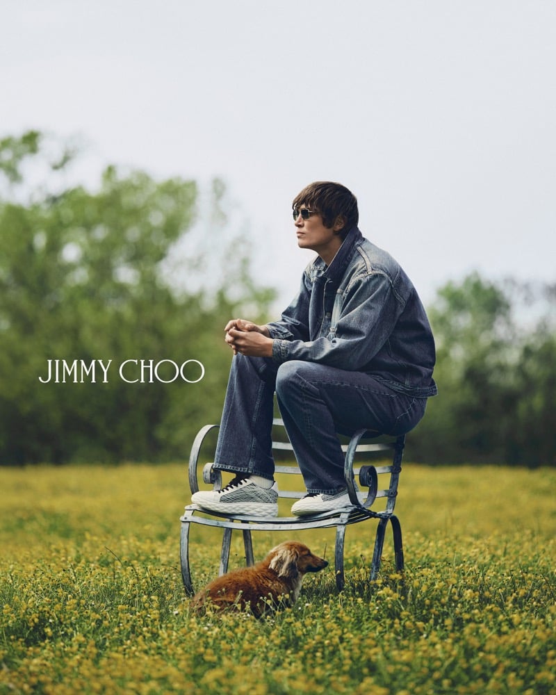 Alfie Nickerson seated on a curling wrought-iron garden chair in a buttercup-filled meadow with his dachshund Wolfie in the grass below, wearing dark sunglasses, a faded blue denim jacket, matching mid-wash jeans, and white Jimmy Choo Diamond Maxi Trainers
