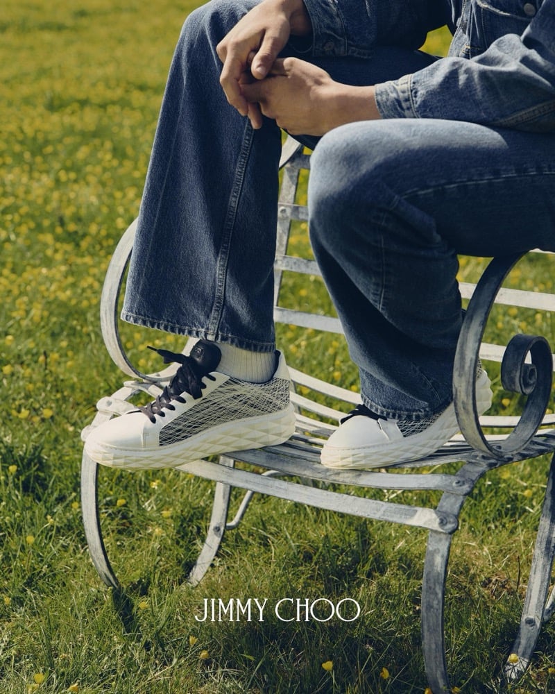 Tight crop on Alfie Nickerson's feet on the wrought-iron chair, showing the Jimmy Choo Diamond Maxi Trainer in woven white leather with a navy lacing system, paired with mid-wash bootcut denim and white ribbed socks