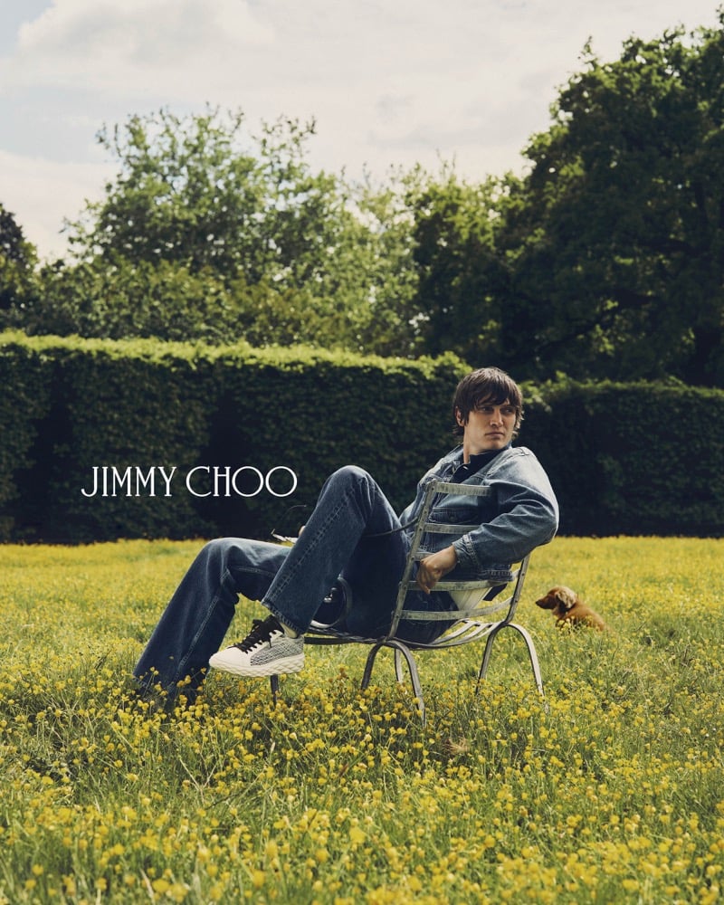 Alfie Nickerson reclined back on the wrought-iron chair in the middle of the buttercup meadow with his dachshund Wolfie standing in the grass behind him, dressed in a denim jacket, matching jeans, and white Jimmy Choo Diamond Maxi Trainers, with a tall green hedge in the distance