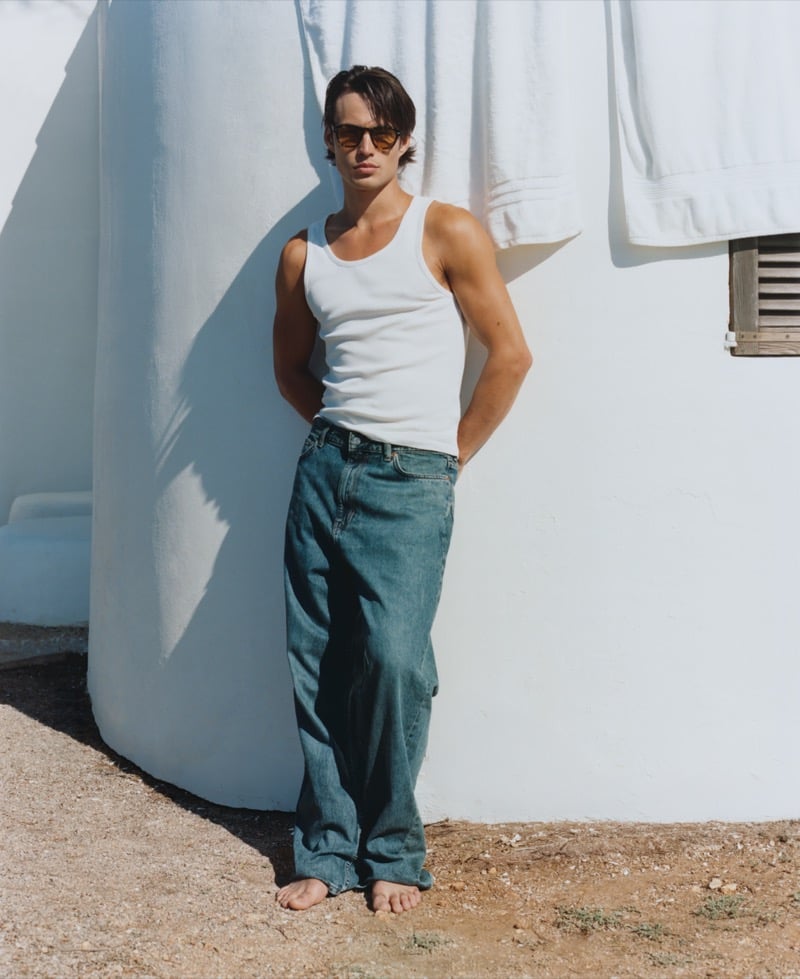 Louis Baines leans barefoot against a white wall wearing Jimmy Fairly's The Headlight yellow tortoiseshell sunglasses with a white ribbed tank top and dark wash baggy jeans
