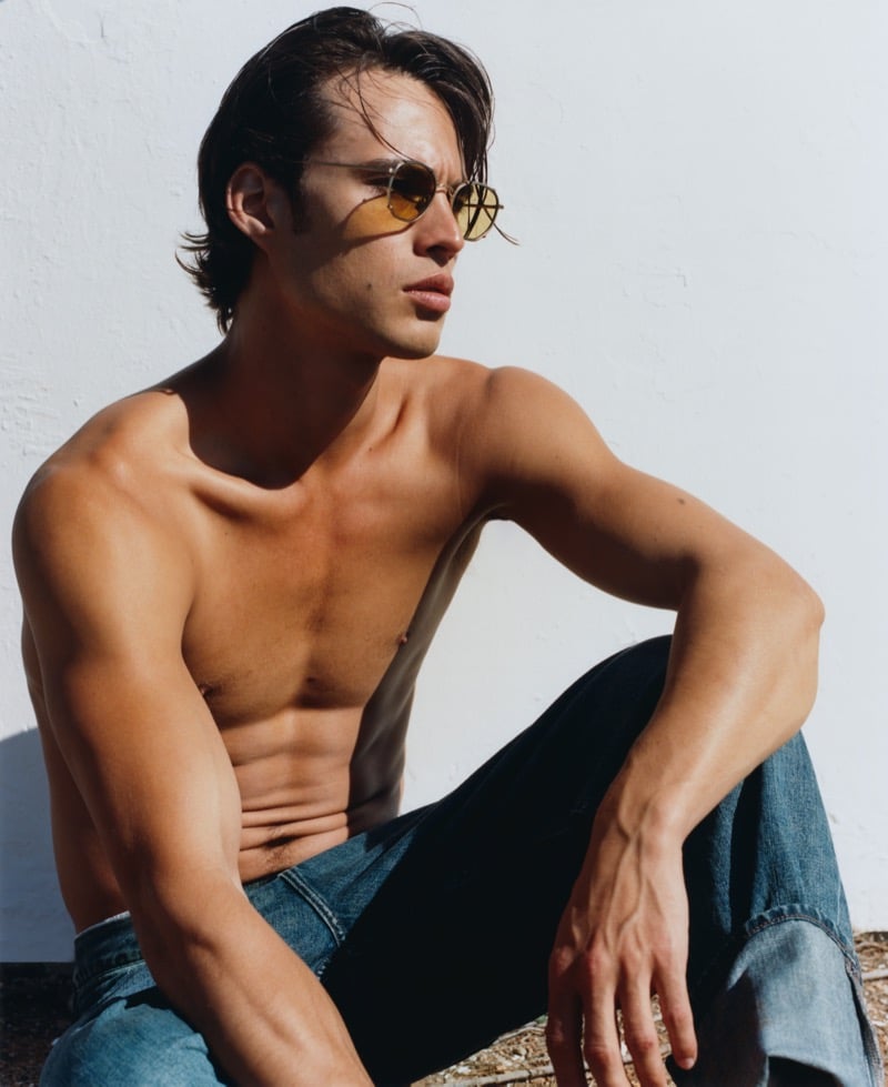 Louis Baines sits shirtless against a white wall in Jimmy Fairly's The Wave round gold-frame sunglasses with yellow lenses and dark wash jeans
