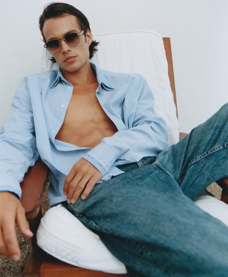 Louis Baines reclines in a deck chair wearing Jimmy Fairly's The Wax brown tortoiseshell sunglasses, an open blue and white striped shirt, light wash jeans, and white sneakers