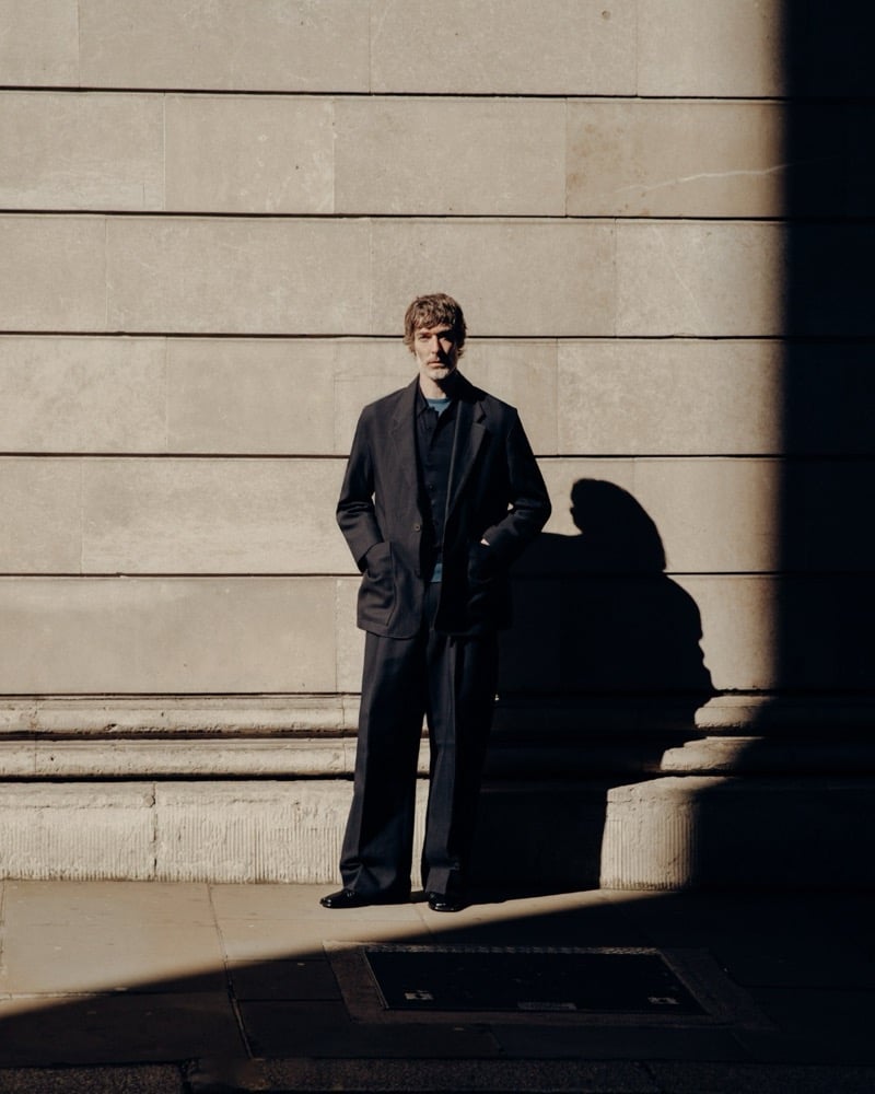 Richard Biedul stands in sunlight and shadow against a neoclassical stone wall in a charcoal wide-shouldered suit worn over the navy John Smedley Adrian polo, the suit jacket open over wide-leg trousers and dark loafers