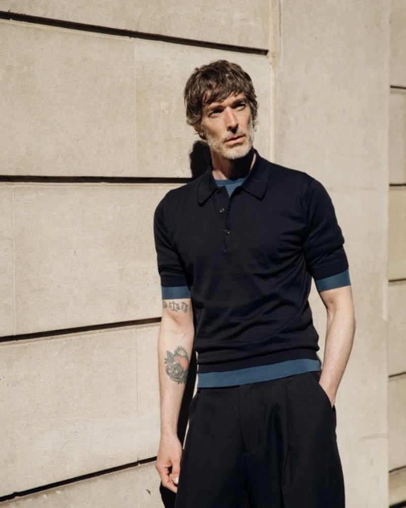 Richard Biedul photographed against a sunlit limestone wall in the John Smedley Adrian knitted polo, layered with a blue top, paired with black pleated wool trousers