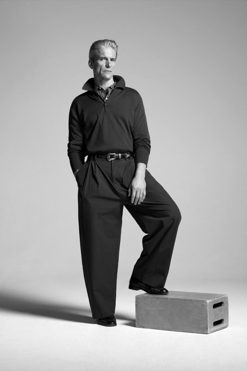 Felix Friedman models the John Smedley Finchley Sea Island cotton long-sleeve polo over a checked shirt with wide-leg trousers, foot resting on an apple box